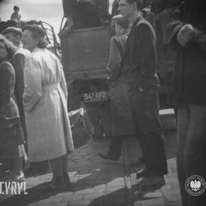 kilka kobiet i mężczyzn zgromadzonych wokół samochodów ciężarowych, z tego dwie kobiety trzymają w rękach teczki