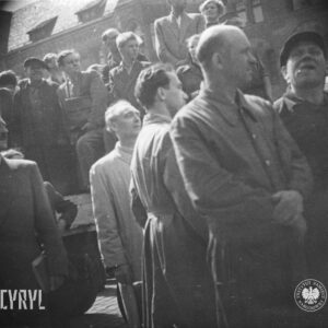 na pierwszym planie kilku mężczyzn, część w roboczych kombinezonach, w głębi grupa mężczyzn stoi na pace samochodu ciężarowego