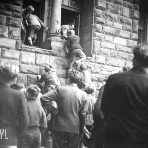 grupa mężczyzn i kilku chłopców stoi przed ścianą budynku z bloków kamiennych, kilku mężczyzn z pomocą innych wchodzi przez otwarte okna do budynku