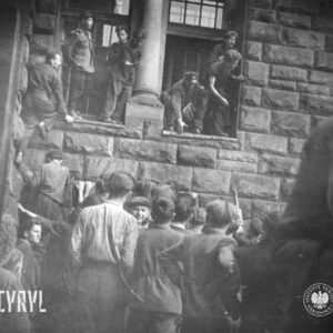 grupa mężczyzn i kilku chłopców stoi przed ścianą budynku z bloków kamiennych, kilku mężczyzn z pomocą innych wchodzi przez otwarte okna do budynku