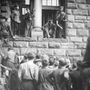 grupa mężczyzn i kilku chłopców stoi przed ścianą budynku z bloków kamiennych, kilku mężczyzn z pomocą innych wchodzi przez otwarte okna do budynku