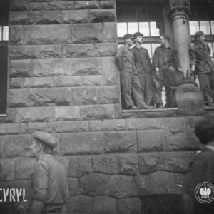 fragment ściany budynku z bloków kamiennych, na parapecie okna na parterowej kondygnacji stoi czterech mężczyzn