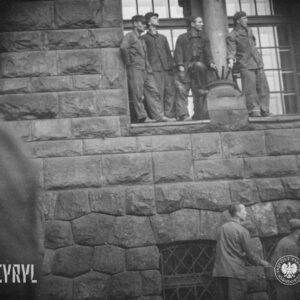 fragment ściany budynku z bloków kamiennych, na parapecie okna na parterowej kondygnacji stoi czterech mężczyzn