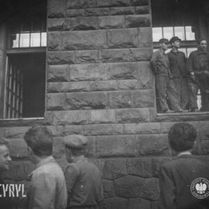 fragment ściany budynku z bloków kamiennych, na parapecie okna na parterowej kondygnacji stoi czterech mężczyzn