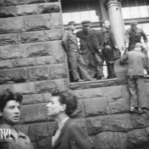 fragment ściany budynku z bloków kamiennych, na parapecie okna na parterowej kondygnacji stoi kilku mężczyzn, jeden mężczyzna z pomocą stojących wchodzi na parapet