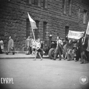 duża grupa ludzi idzie ulicą, na czele trzech młodych mężczyzn idzie z transparentem, po obu ich stronach idą chłopiec i mężczyzną z flagami, w głębi ściana masywnego gmachu