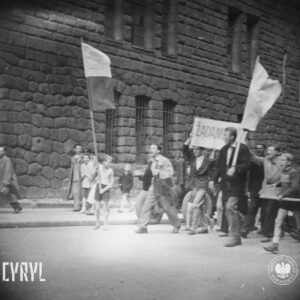 duża grupa ludzi idzie ulicą, na czele trzech młodych mężczyzn idzie z transparentem, po obu ich stronach idą chłopiec i mężczyzną z flagami, w głębi ściana masywnego gmachu