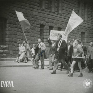 duża grupa ludzi idzie ulicą, na czele trzech młodych mężczyzn idzie z transparentem, po obu ich stronach idą chłopiec i mężczyzną z flagami, w głębi ściana masywnego gmachu