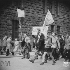 duża grupa ludzi idzie ulicą, na czele idzie mężczyzna z flagą, towarzyszy mu chłopiec, w głębi ściana masywnego gmachu