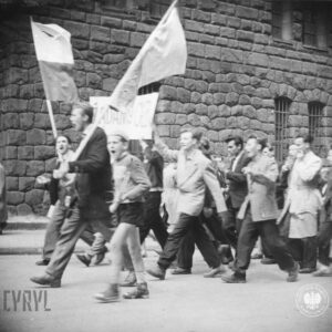 grupa ludzi idzie ulicą, na jej czele mężczyzna z flagą, towarzyszy mu chłopiec, w głębi ściana masywnego gmachu