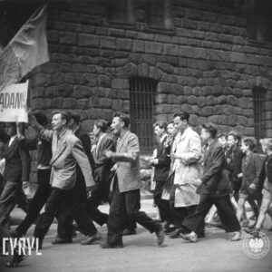 duża grupa ludzi idzie ulicą, na czele trzech młodych mężczyzn idzie z transparentem, na końcu grupy idą chłopcy, w głębi ściana masywnego gmachu