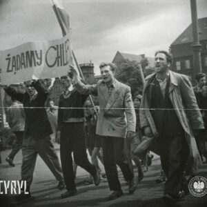 grupa młodych mężczyzn idzie na czele grupy, trzech z nich trzyma nad głowami transparent z hasłem, za nimi idą chłopcy, w głębi budynki
