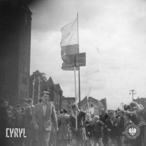 pośrodku kilku chłopców otoczonych grupą dorosłych, jeden z chłopców trzyma tablicę przymocowaną do kija, za nim kilka flag