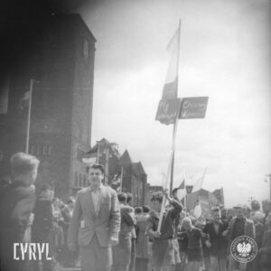 pośrodku kilku chłopców otoczonych grupą dorosłych, jeden z chłopców trzyma tablicę przymocowaną do kija, za nim kilka flag