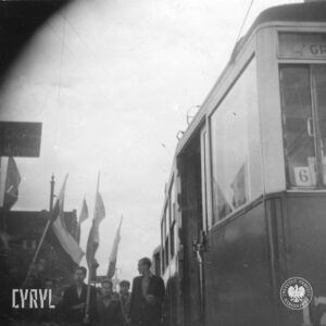 grupa chłopców i kilku młodych mężczyzn, część trzyma flagi, z prawej tramwaj