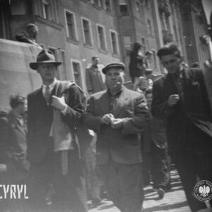 grupa ludzi idących ulicą, jeden z mężczyzn w garniturze, płaszczem przewieszonym przez ramię i w kapeluszu, w głębi budynki
