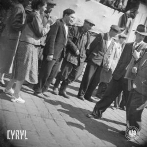 grupa ludzi idących ulicą, jeden z mężczyzn w garniturze, płaszczem przewieszonym przez ramię i w kapeluszu, na chodniki maszerujących obserwują ludzie, część w roboczych kombinezonach, jedna z kobiet klaszcze, w głębi budynki