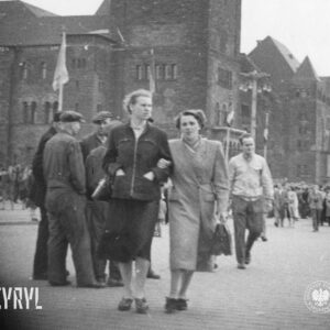 na pierwszym planie dwie kobiety idą ulicą, trzymając się pod rękę, w głębi bardzo duże zgromadzenie ludzi i potężny budynek z wysoką wieżą