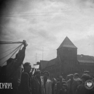 grupa kilkunastu osób ujętych na zdjęciu od pasa w górę, w głębi potężny budynek z wieżą
