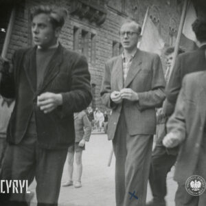 na pierwszym planie dwóch mężczyzn, w głębi pod ścianą potężnego budynku grupa ludzi
