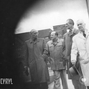 czterej starsi mężczyźni idą ulicą, w głębi okazały budynek