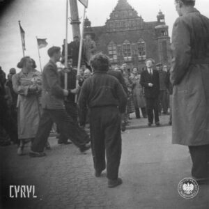 duża grupa osób na placu, pośrodku dwaj mężczyźni niosą transparent, w głębi okazały budynek