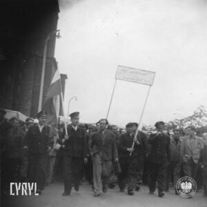 duża grupa ludzi idzie ulicą, na przodzie dwóch mężczyzn trzyma transparent, część ma w rękach flagi, trzech mężczyzn w pierwszym rzędzie w mundurach