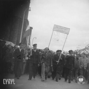 duża grupa ludzi idzie ulicą, na przodzie dwóch mężczyzn trzyma transparent, część ma w rękach flagi, trzech mężczyzn w pierwszym rzędzie w mundurach