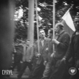 grupa ludzi idzie ulicą, jeden z mężczyzn trzyma flagę, w głębi drzewa