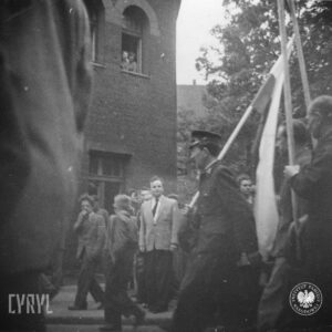 duża grupa ludzi, pośrodku mężczyzna w mundurze z flagą, w głębi fragment budynku