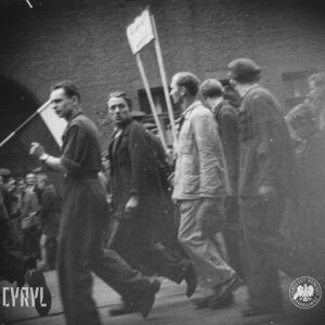 grupa mężczyzn, część w kombinezonach roboczych, idzie ulicą, niosą transparent i flagę, w głębi fragment elewacji budynku