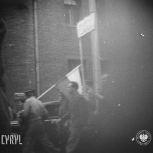 bardzo niewyraźne zdjęcie, kilku mężczyzn, jeden niesie flagę, w głębi fragment elewacji budynku