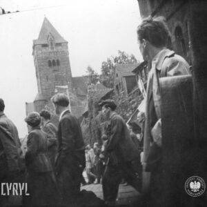 grupa ludzi idzie ulicą, w głębi potężny budynek z wysoką wieżą