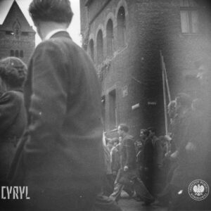 grupa ludzi idzie ulicą, w głębi fragment potężnego budynku z wieżą