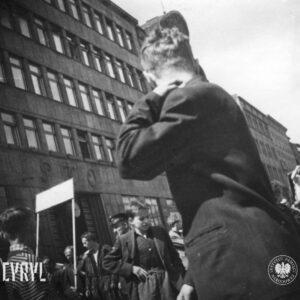 grupa ludzi, na pierwszym planie kilkunastoletni chłopcy, przed kilkupiętrowym modernistycznym budynkiem