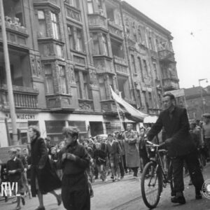 duża grupa dzieci idzie ulicą, wśród nich mężczyzna z flagą, na pierwszym planie mężczyzna prowadzi rower, w głębi kamienice