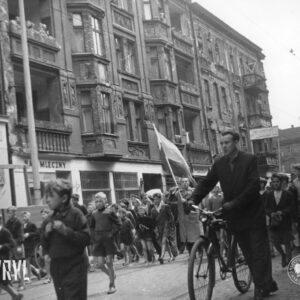 duża grupa dzieci idzie ulicą, wśród nich mężczyzna z flagą, na pierwszym planie mężczyzna prowadzi rower, w głębi kamienice