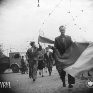 grupa ludzi z flagami na skrzyżowaniu brukowanych ulic z torami tramwajowymi, kilka osób biegnie, między ludźmi samochody ciężarowe