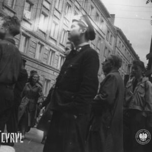 grupa mężczyzn stoi na ulicy, w centrum mężczyzna w mundurze, w głębi kamienice