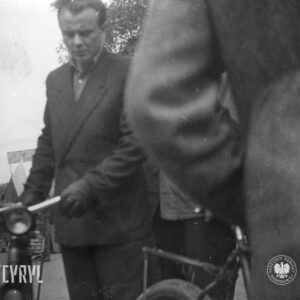 mężczyzna w garniturze prowadzi rower przysłonięty innym mężczyzną sfotografowanym tyłem