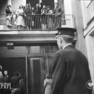 grupa ludzi, kilka osób coś krzyczy, kilka macha rękoma, stoi na balkonie nad wejściem do budynku