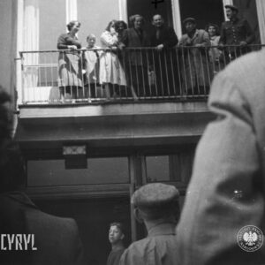 grupa ludzi, kilka osób coś krzyczy, kilka macha rękoma, stoi na balkonie nad wejściem do budynku