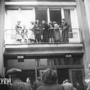 grupa ludzi, kilka osób coś krzyczy, kilka macha rękoma, stoi na balkonie nad wejściem do budynku