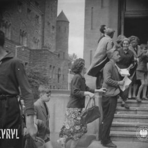 mężczyźni, kobiety i dzieci stoi na schodach prowadzących do budynku, część po nich wchodzi, kilka osób trzyma w rękach arkusze papieru