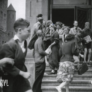 mężczyźni, kobiety i dzieci stoi na schodach prowadzących do budynku, część po nich wchodzi, kilka osób trzyma w rękach arkusze papieru