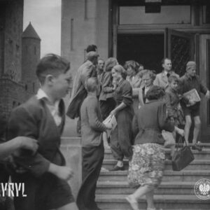 mężczyźni, kobiety i dzieci stoi na schodach prowadzących do budynku, część po nich wchodzi, kilka osób trzyma w rękach arkusze papieru
