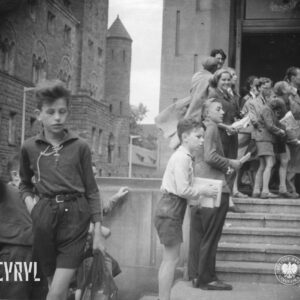 na pierwszym planie chłopiec z siatką i jedną ręką w kieszeni, za nim mężczyźni, kobiety i dzieci, stoją przed wejściem do budynki