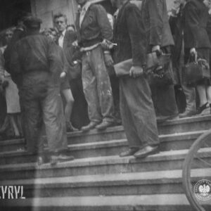 grupa ludzi na schodach przed wejściem do budynku, wśród stojących wyróżnia się mężczyzna w roboczym ubraniu poplamionym jasną farbą lub gipsem