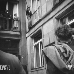 mężczyzna w roboczym ubraniu, poplamionym białą farbą lub gipsem z drewnianym kijem w ręku stoi po zewnętrznej stronie barierki balkonu, przygląda mu się mężczyzna stojący przed budynkiem