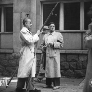 dwóch mężczyzn w prochowcach stoi przed budynkiem, mężczyzna z lewej (oznaczony krzyżykiem) trzyma w rękach drewniany kij